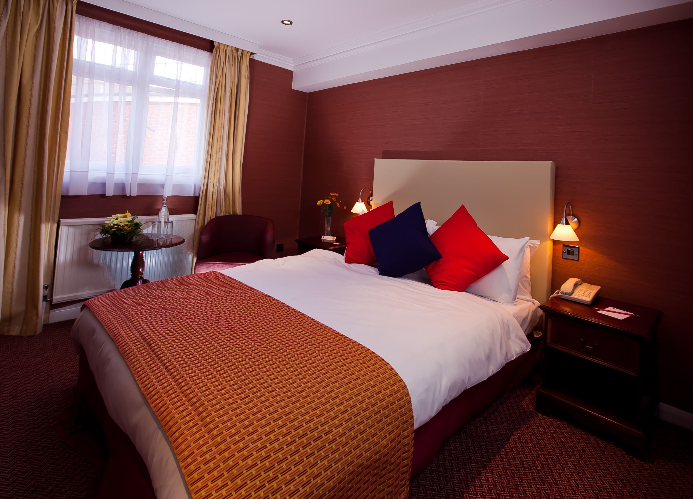 Ein Hotelzimmer mit einem großen Bett, roten und blauen Kissen, einem Nachttisch mit Lampe und einem Sessel am Fenster.
