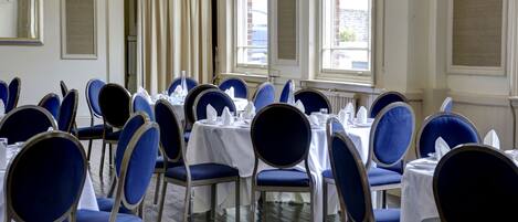 A dining area with round tables and blue chairs, large windows, and a white tablecloth.