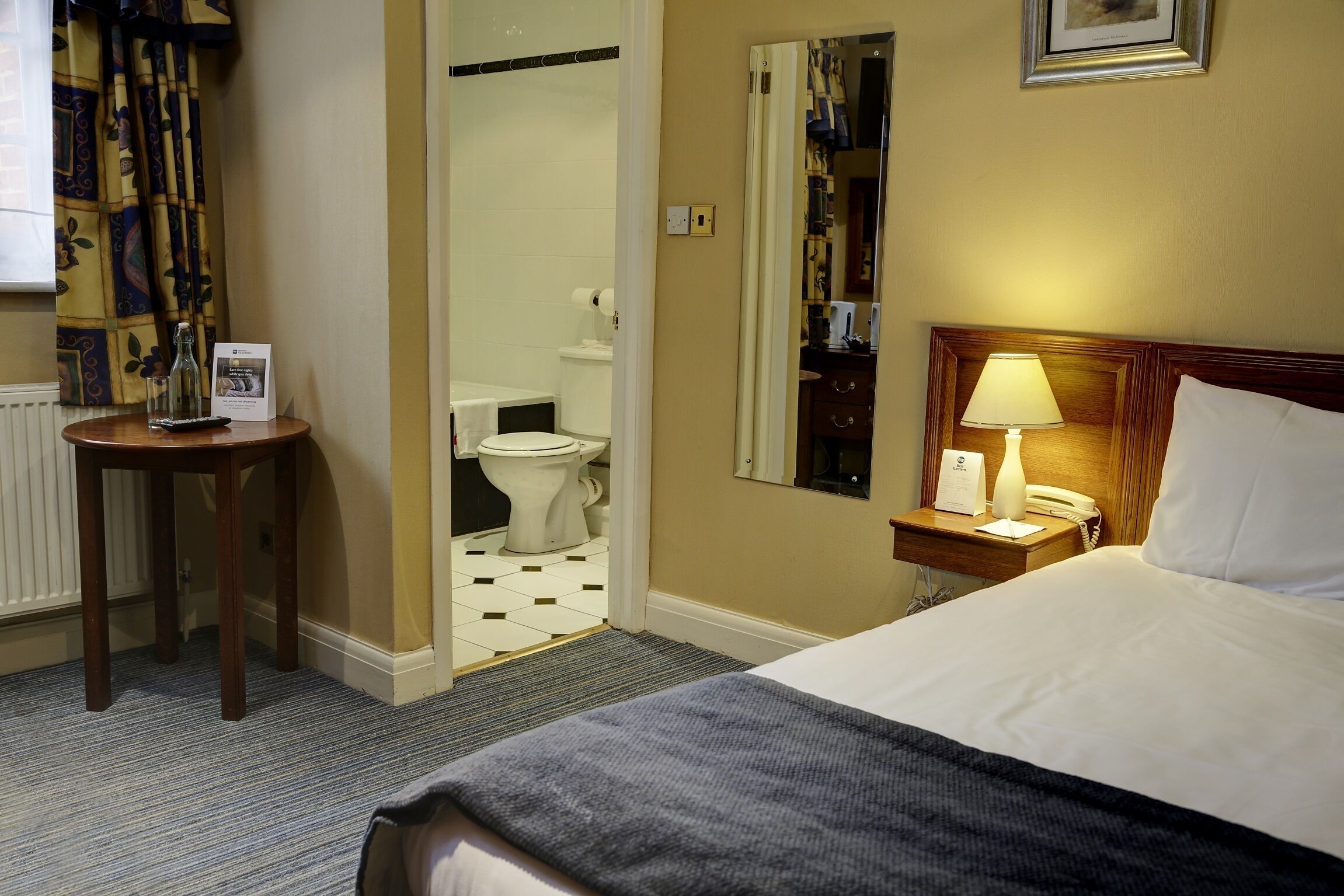 Camera d'albergo con un letto, un comodino con una lampada, un tavolino con un bicchiere e una brochure, uno specchio e vista sul bagno.