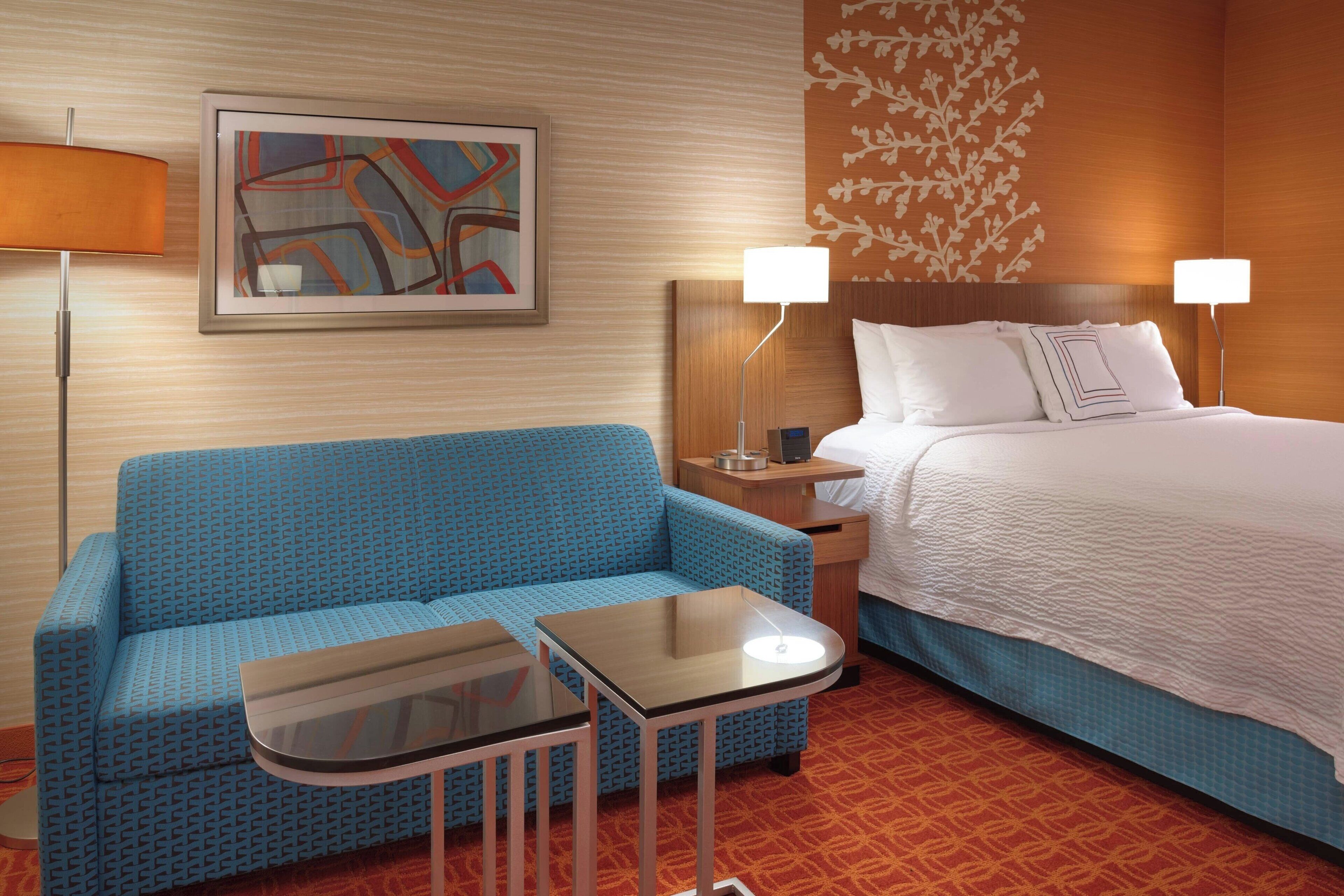 Habitación de hotel con sofá azul, una mesita, cama con ropa de cama blanca y dos lámparas de noche.