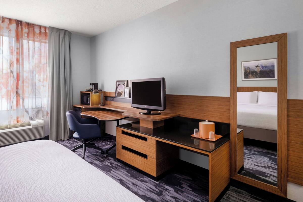 A hotel room with a bed, a desk with a computer, a chair, a mirror, and a window with curtains.