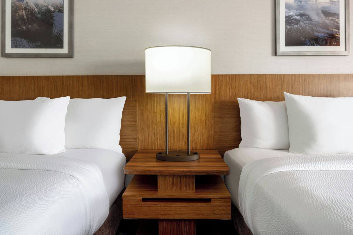 A hotel room with two beds, a wooden nightstand with a lamp, and framed artwork on the wall.