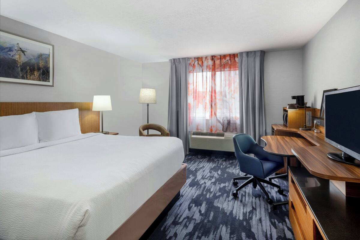 A hotel room with a large bed, a desk with a computer, a chair, a TV, and a window with curtains.