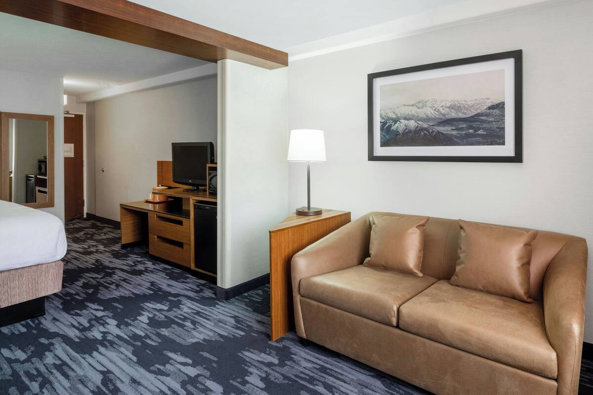 A hotel room with a bed, a sofa, a desk with a lamp, a television, and a framed picture on the wall.