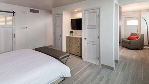 Quarto com cama, um banco, cômoda, televisão e uma cadeira com almofada vermelha.