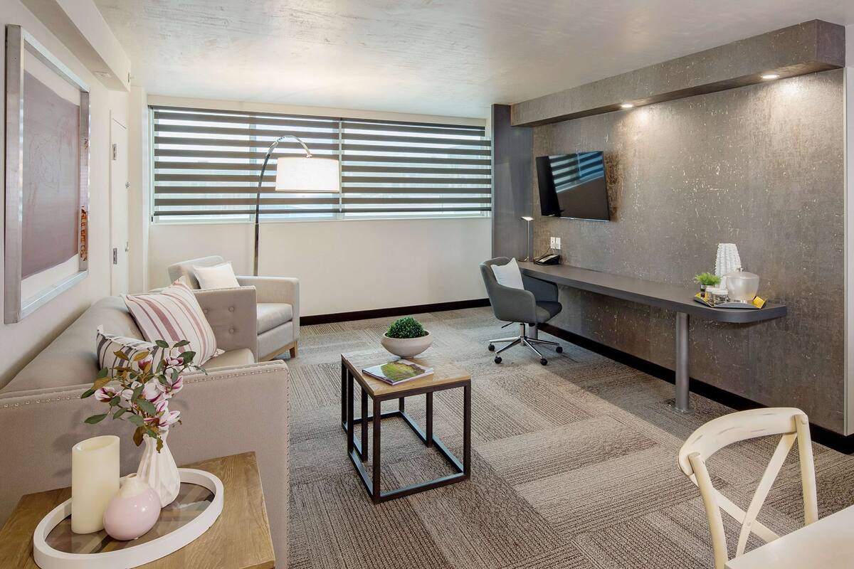 Une chambre d’hôtel moderne comprenant un canapé, une petite table avec un vase et des bougies, un bureau avec une chaise, un téléviseur à écran plat fixé au mur et un lit avec du linge de maison blanc.