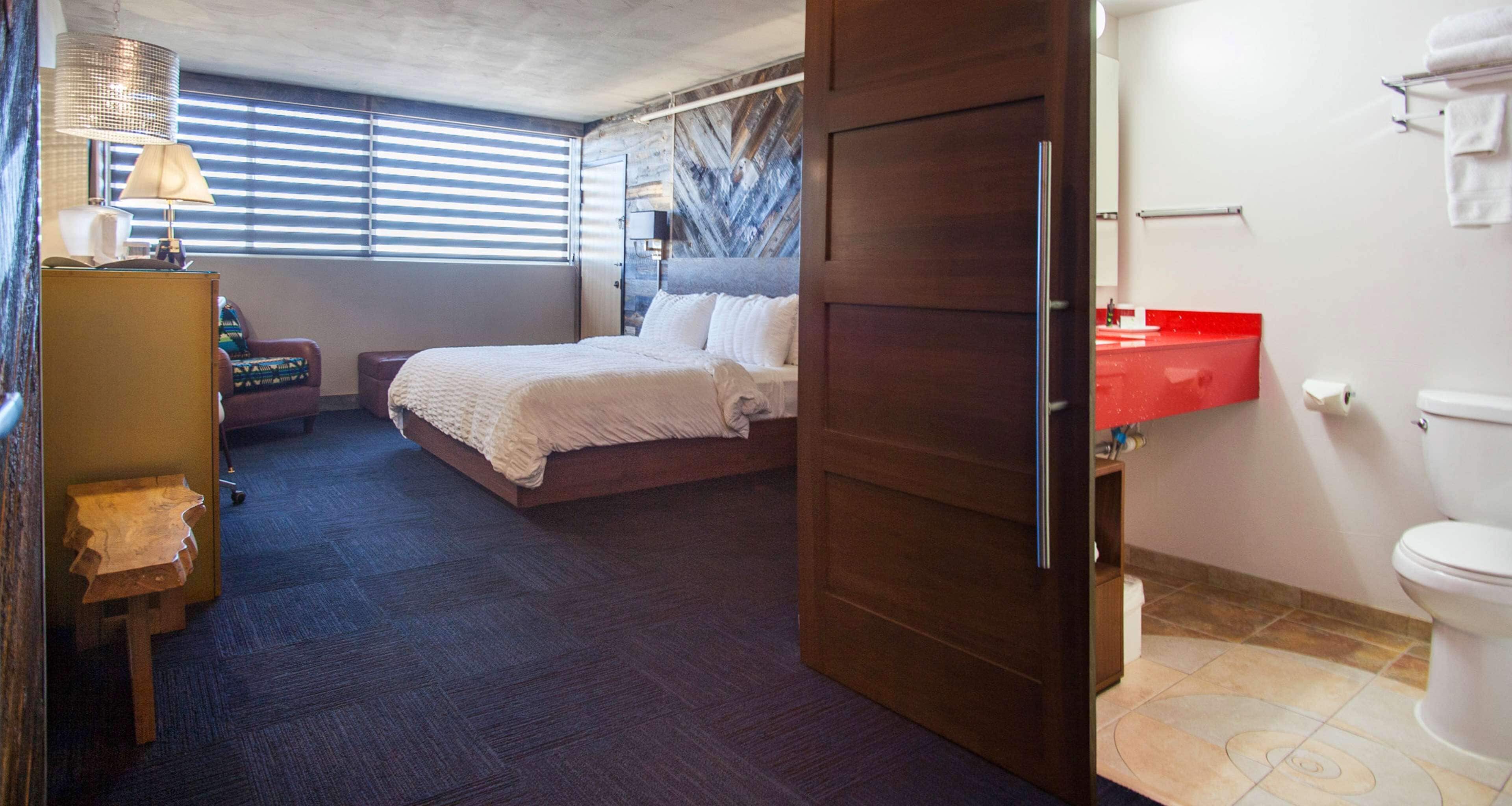 A hotel room with a bed, a desk with a lamp, a chair, a bathroom with a red sink and toilet.