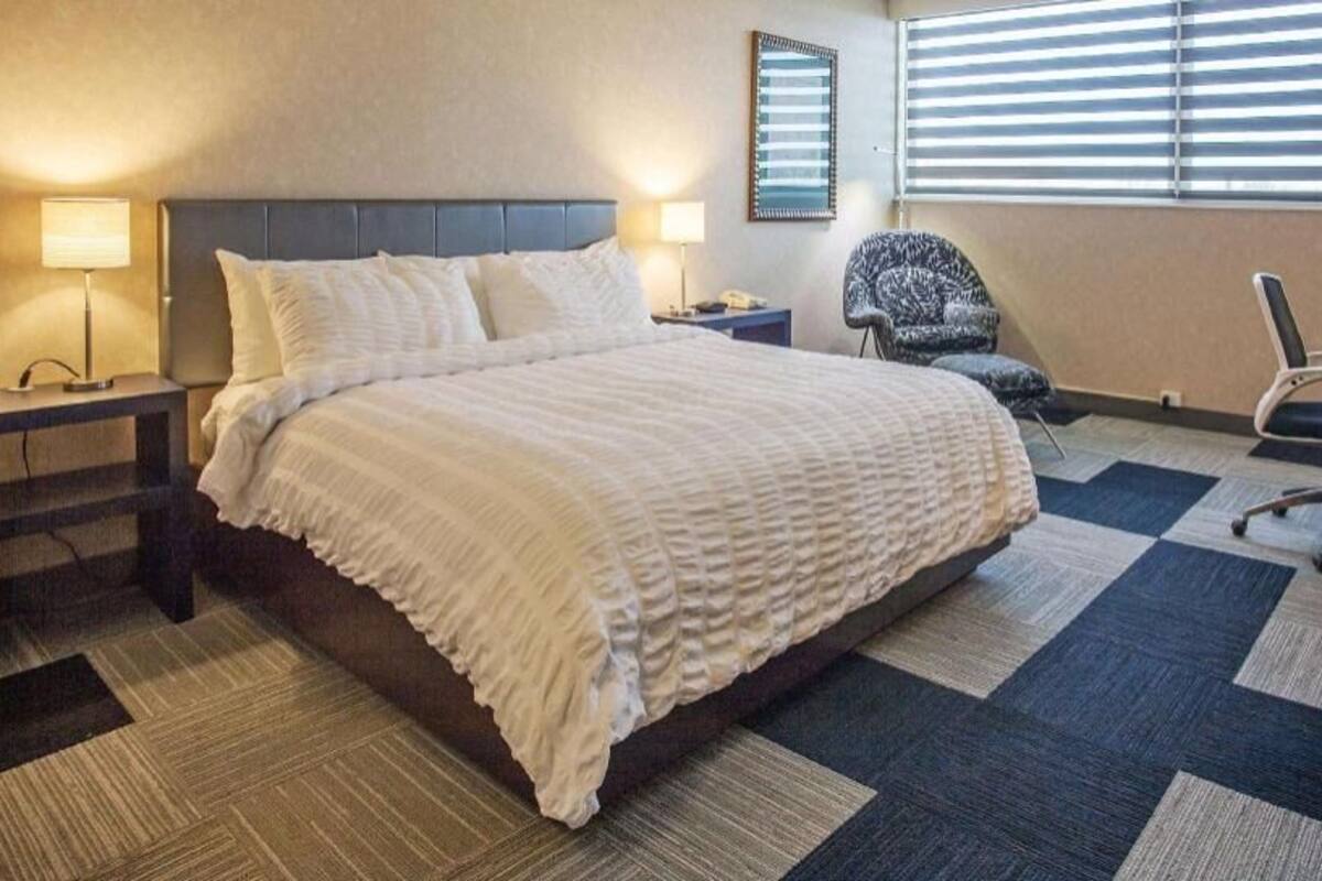 Une chambre d’hôtel avec un grand lit, deux tables de chevet avec des lampes, une chaise, un bureau et une fenêtre avec des stores.