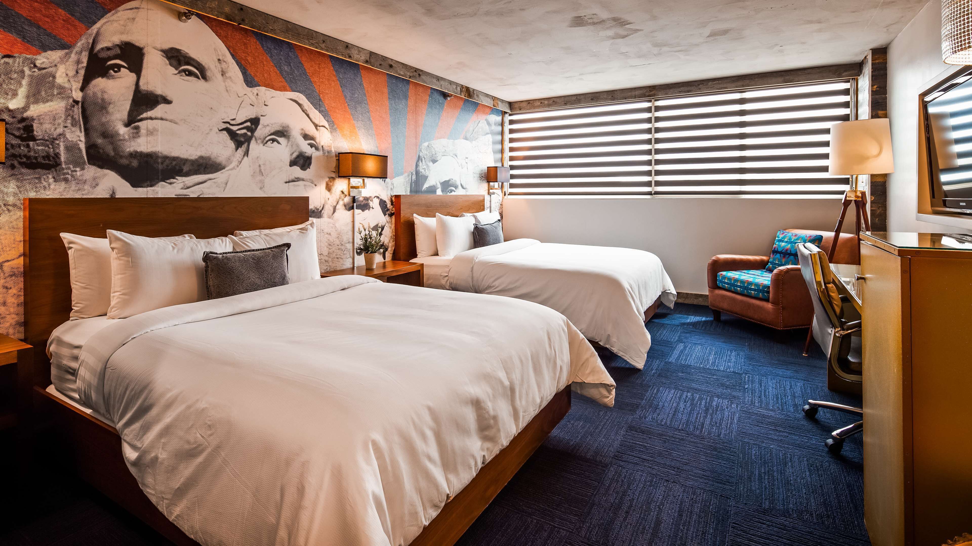 Habitación de hotel con dos camas, un mural grande en la pared, un escritorio con lámpara y una ventana con persianas.
