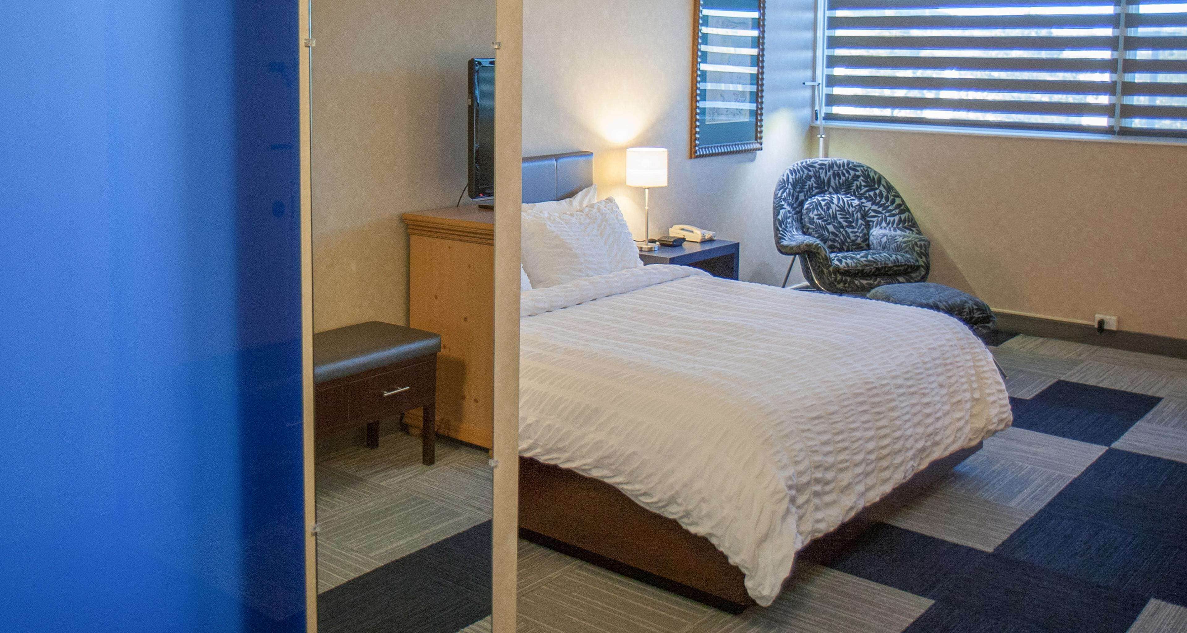 Une chambre d’hôtel comprenant un lit, une table de chevet, une lampe, une chaise et une fenêtre avec des stores.