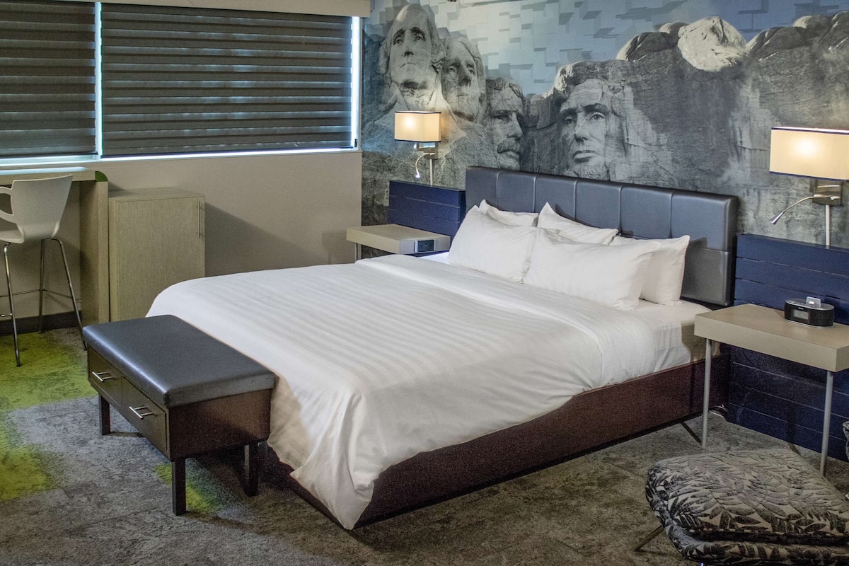 Une chambre d’hôtel avec un grand lit, deux tables de chevet avec des lampes, un bureau avec une chaise et un fauteuil à motifs.