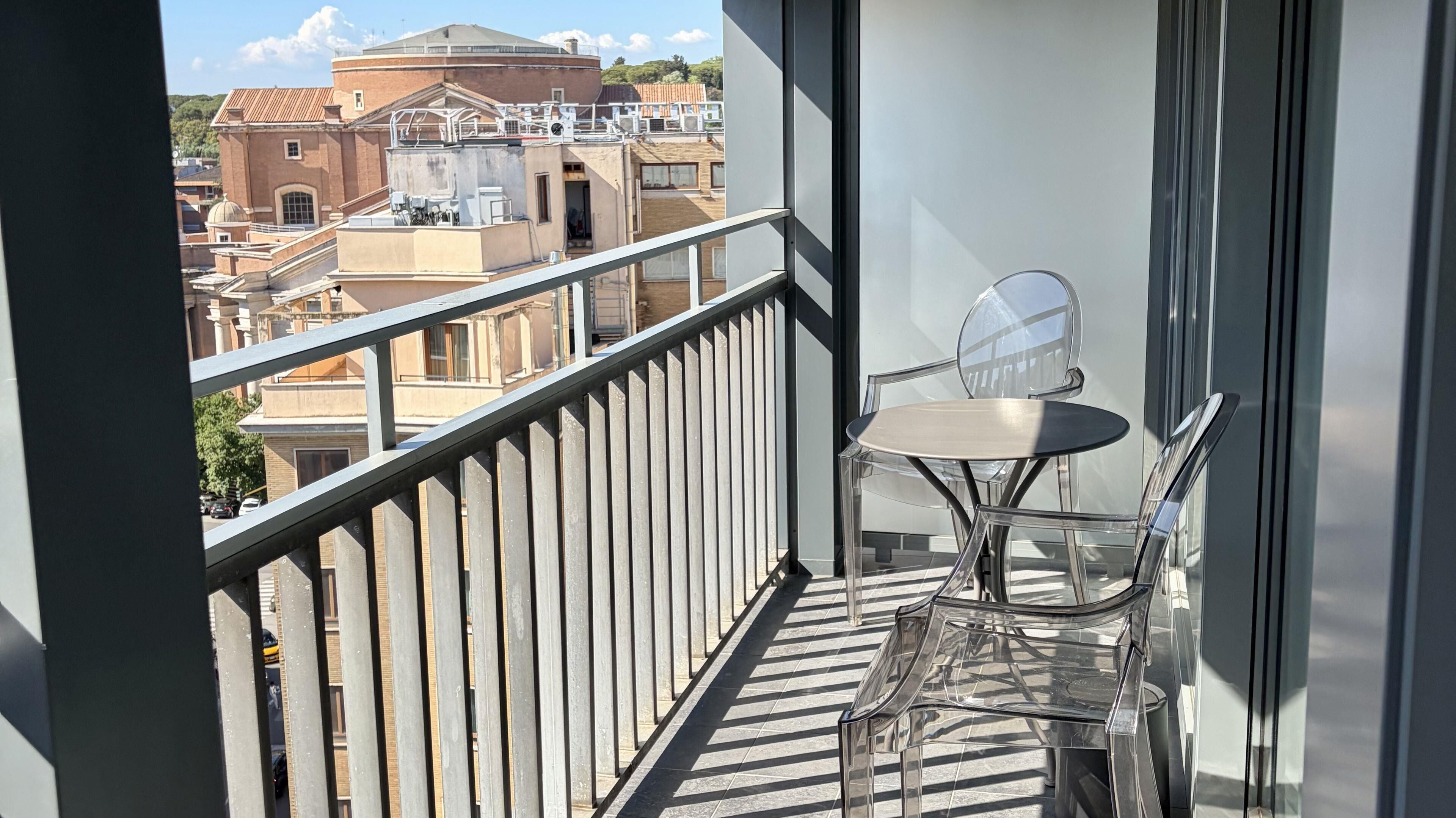 A balcony with a glass table and chairs, a metal railing, and a view of buildings in the background.
