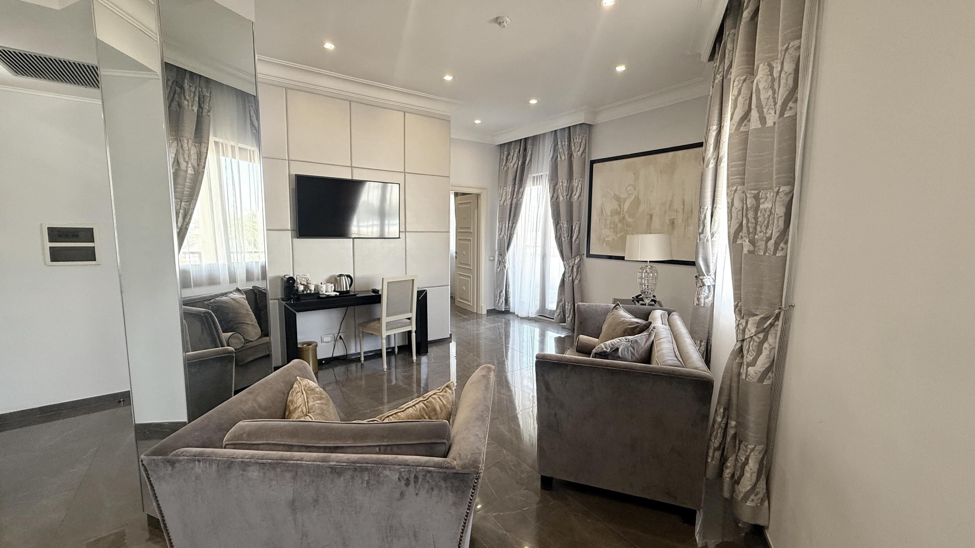 A modern living room with a flat-screen TV, two sofas, a dining area with a table and chairs, and a large window with curtains.