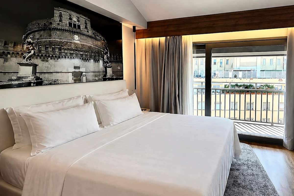 A hotel room with a large bed, a view of a cityscape, a balcony with a railing, and a statue on the wall.