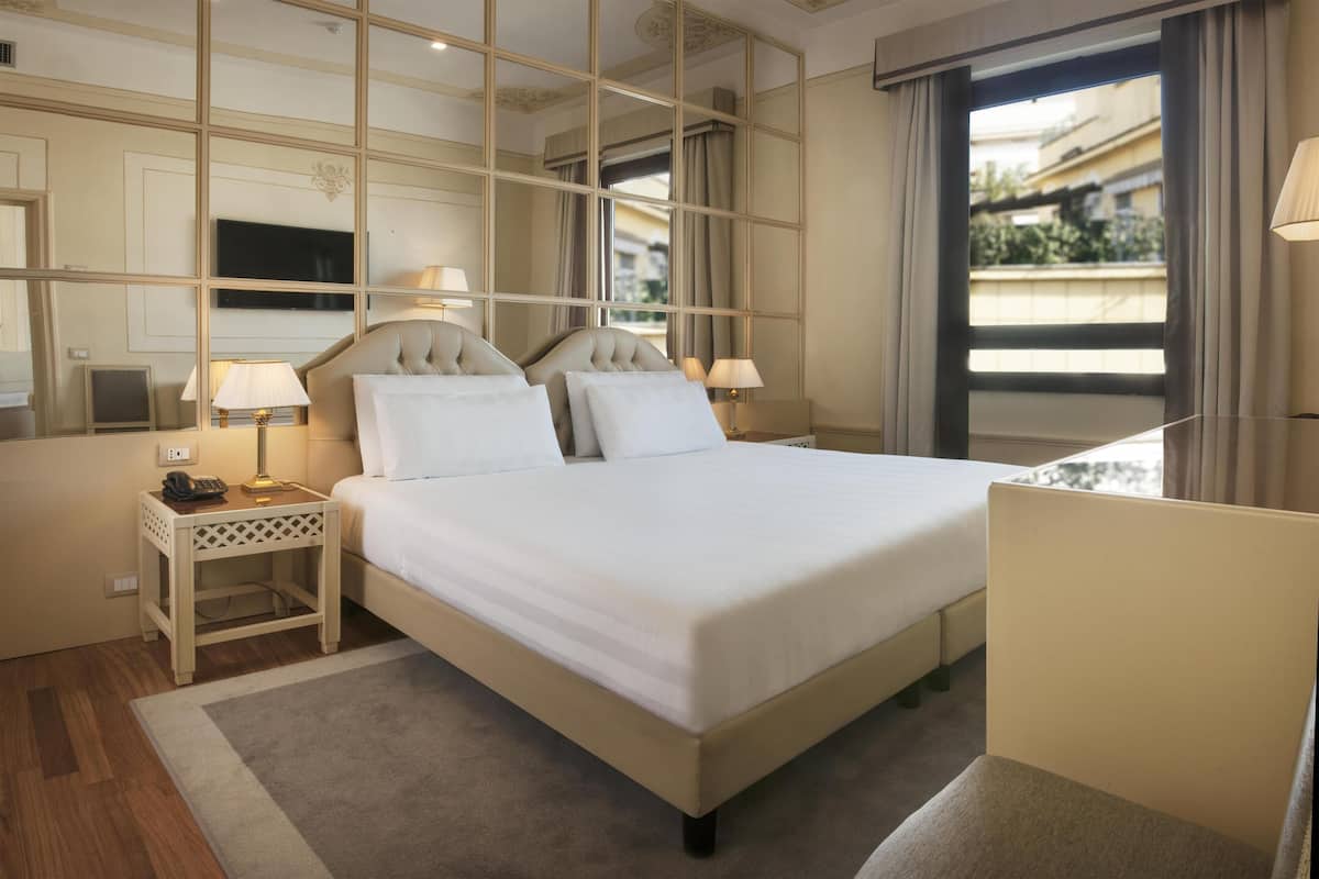 A hotel room with a large bed, bedside lamps, a small table, and a mirror.