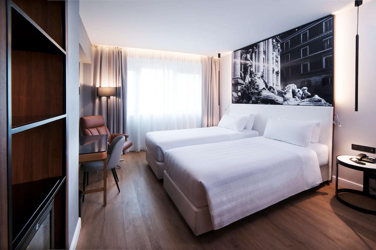 A hotel room with two beds, a desk, a chair, a lamp, and a large black and white photograph on the wall.