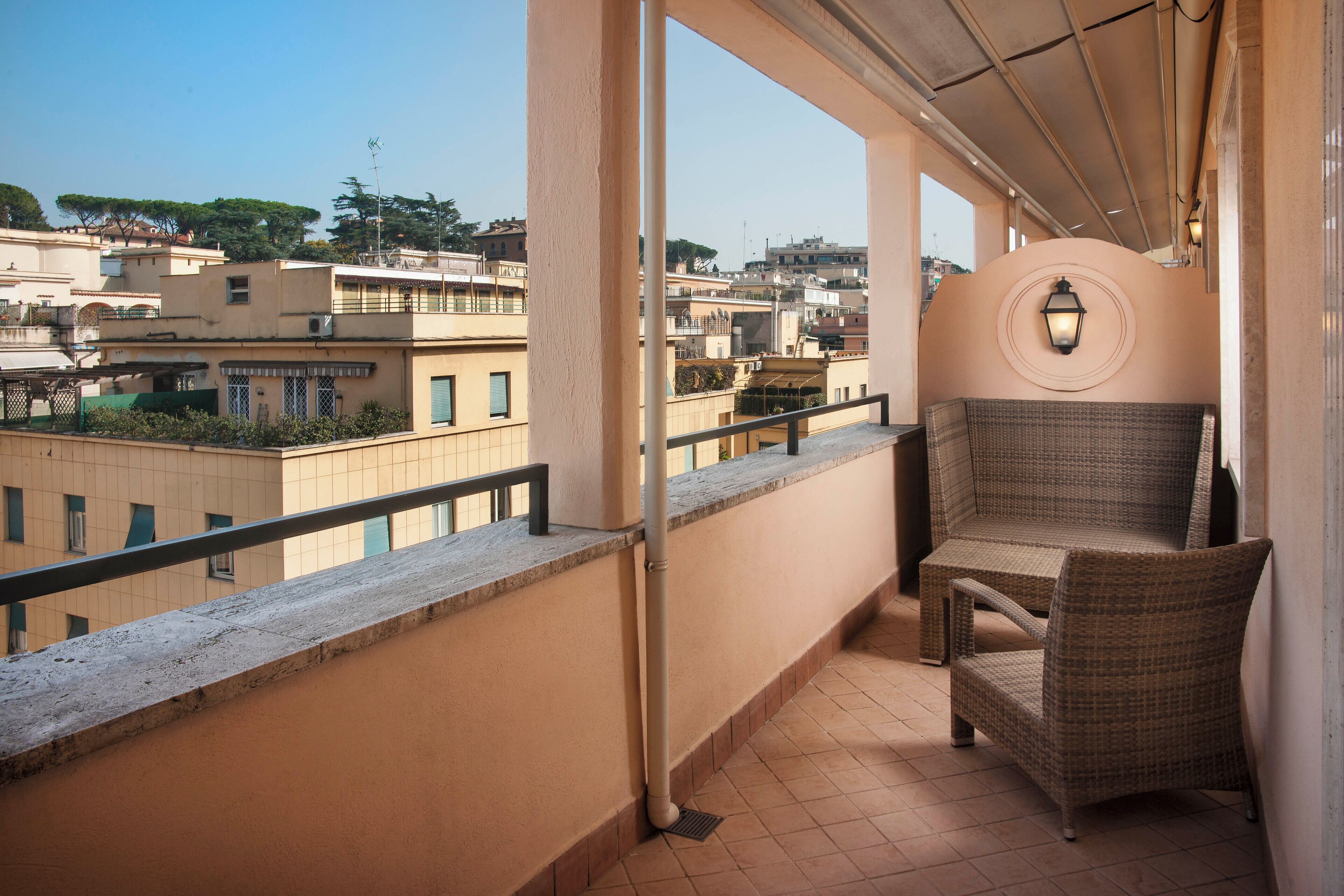 A balcony with a wicker chair, a view of surrounding buildings, and a wall-mounted light fixture.