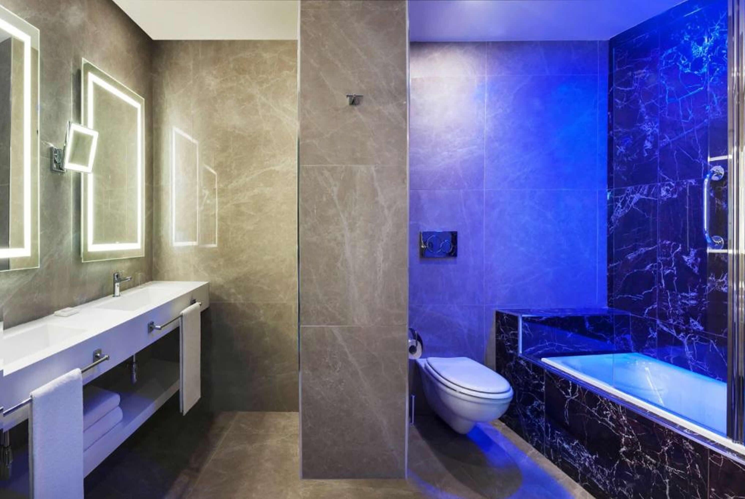 A modern bathroom with a large mirror, a white sink, a toilet, and a bathtub with a marble surround.