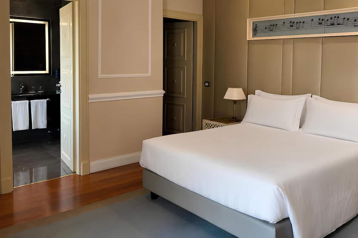 A neatly made bed with white linens, a headboard with a music-themed design, a bedside table with a lamp, and a door leading to a bathroom.