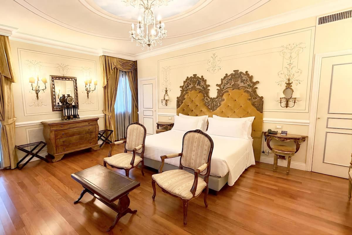 A classic hotel room with a large bed, two chairs, a wooden dresser, a small table, and a chandelier.