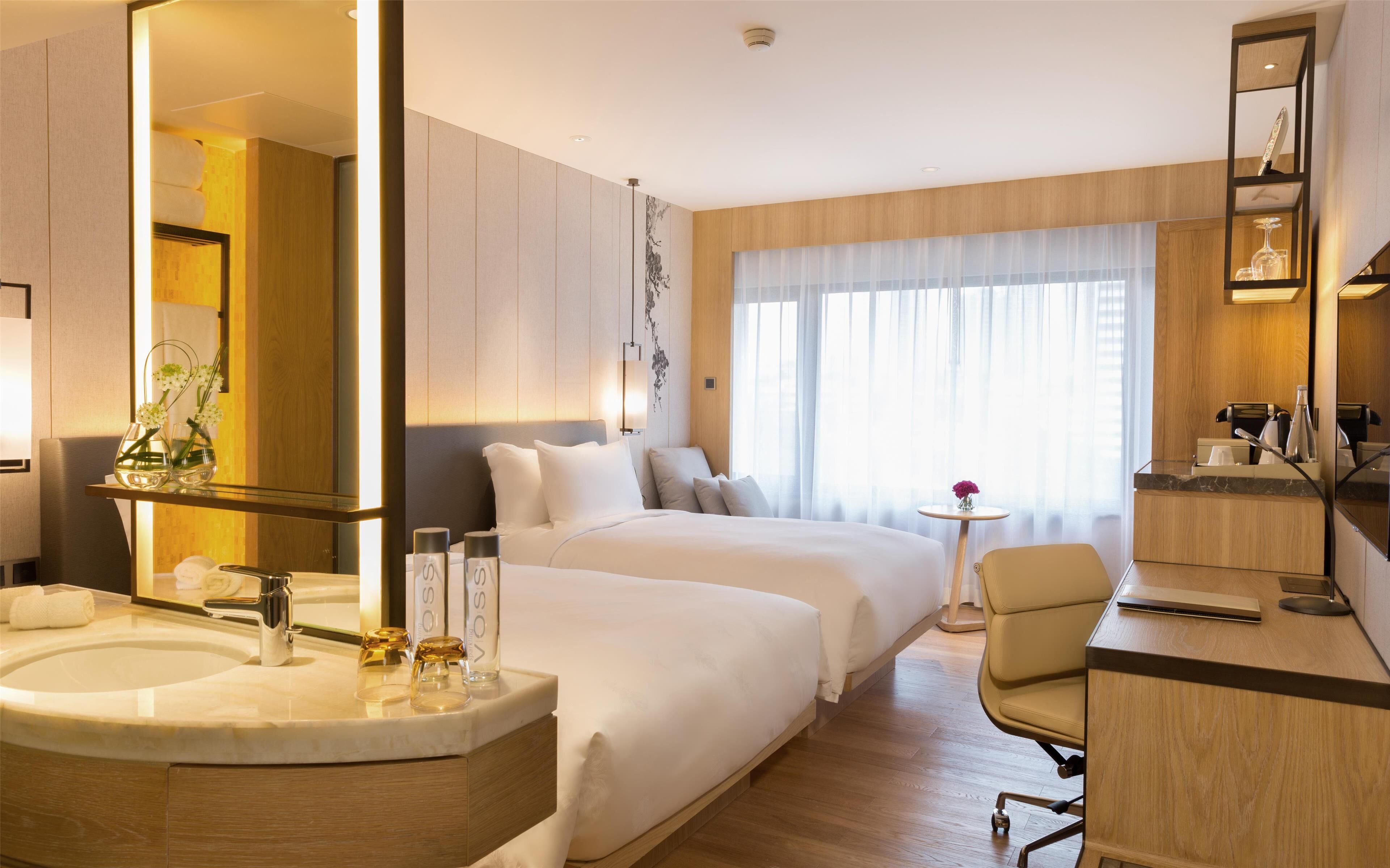 A hotel room with a large bed, a desk, a chair, a bathroom with a sink and mirror, and a large window with curtains.