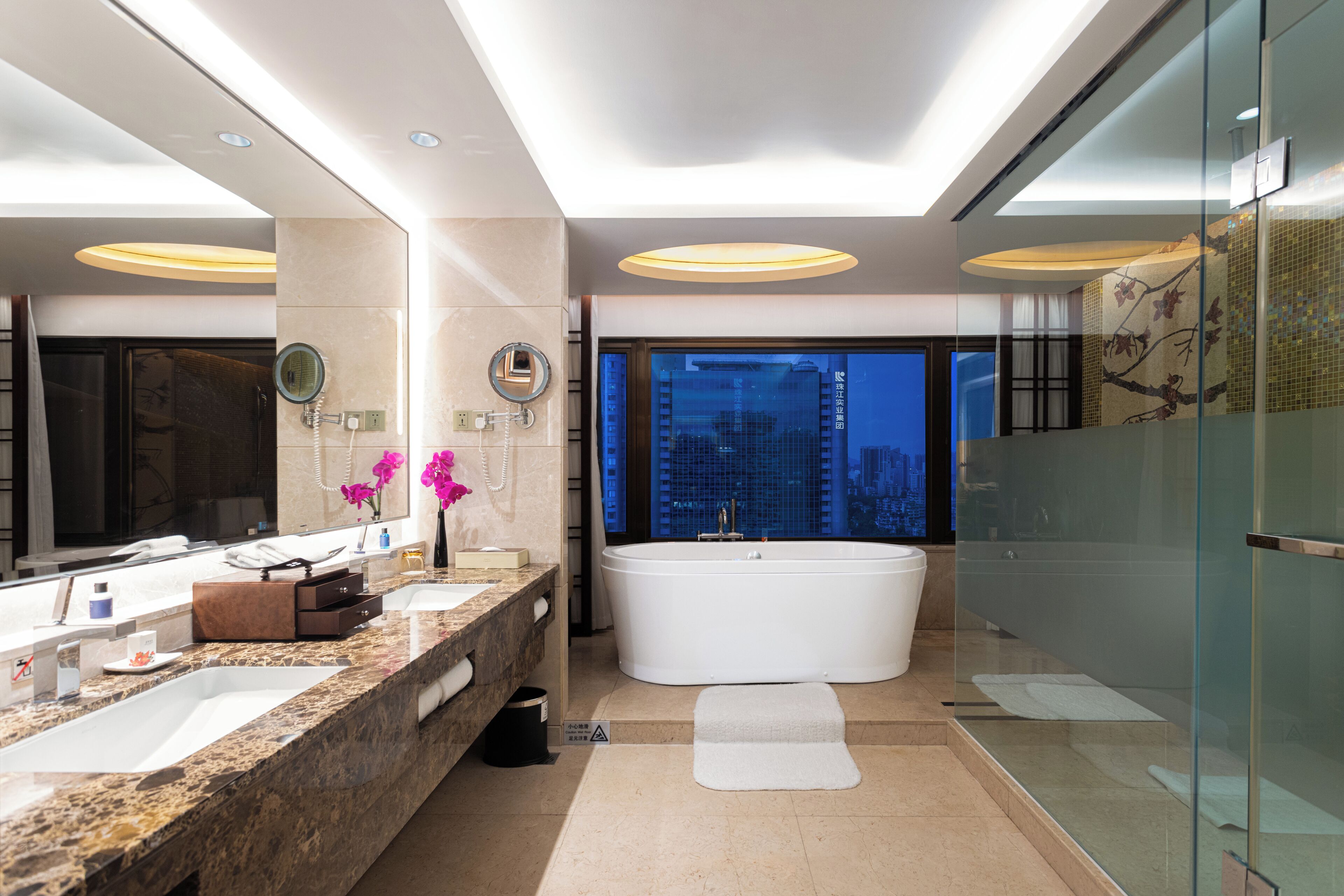 Une salle de bain moderne dotée d’une grande baignoire, de deux vasques, d’un meuble-lavabo avec miroir et offrant une vue sur le paysage urbain.