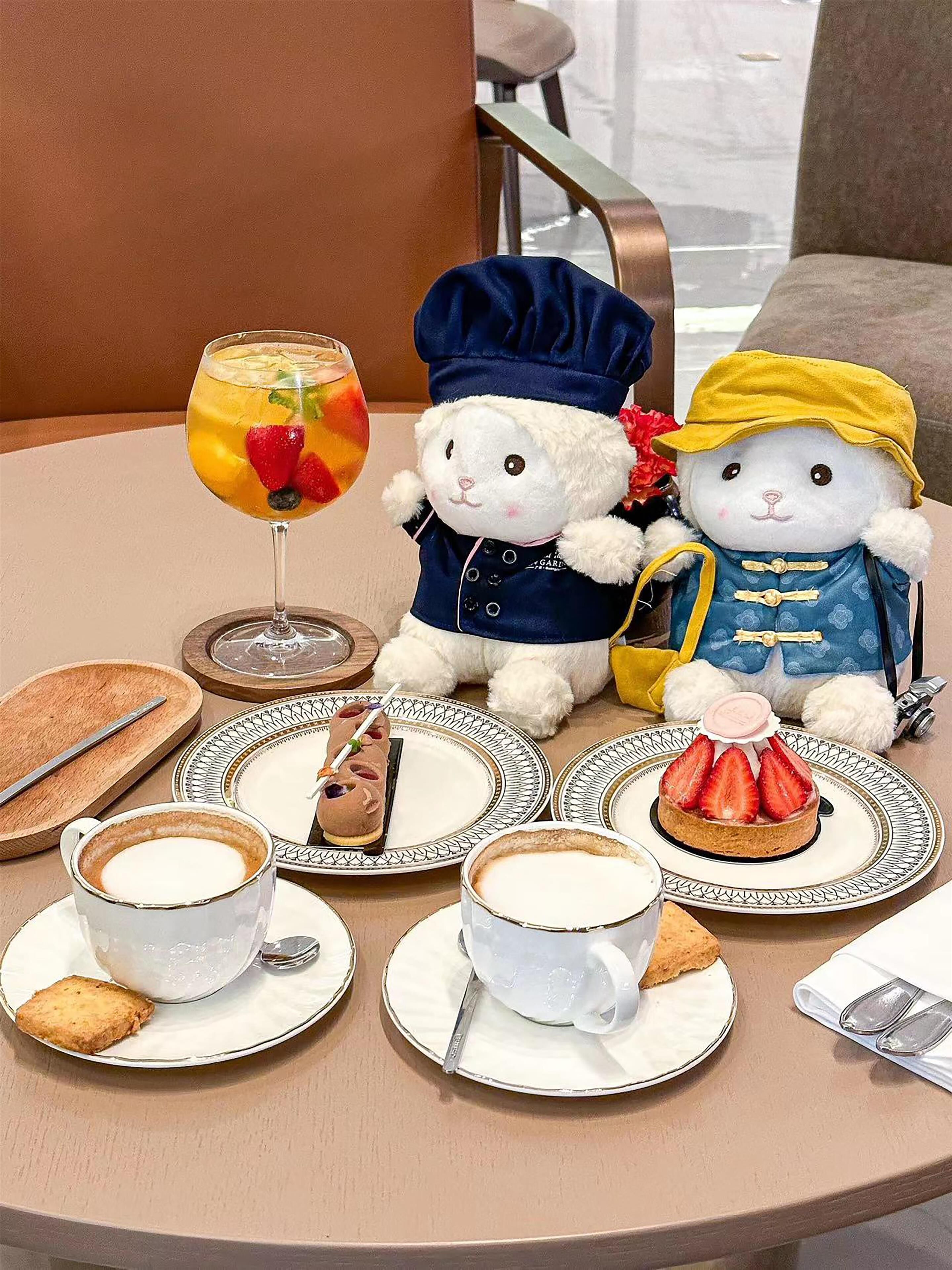 Una mesa dispuesta con un vaso de jugo de frutas, dos platos con postres, una taza de café y dos peluches.