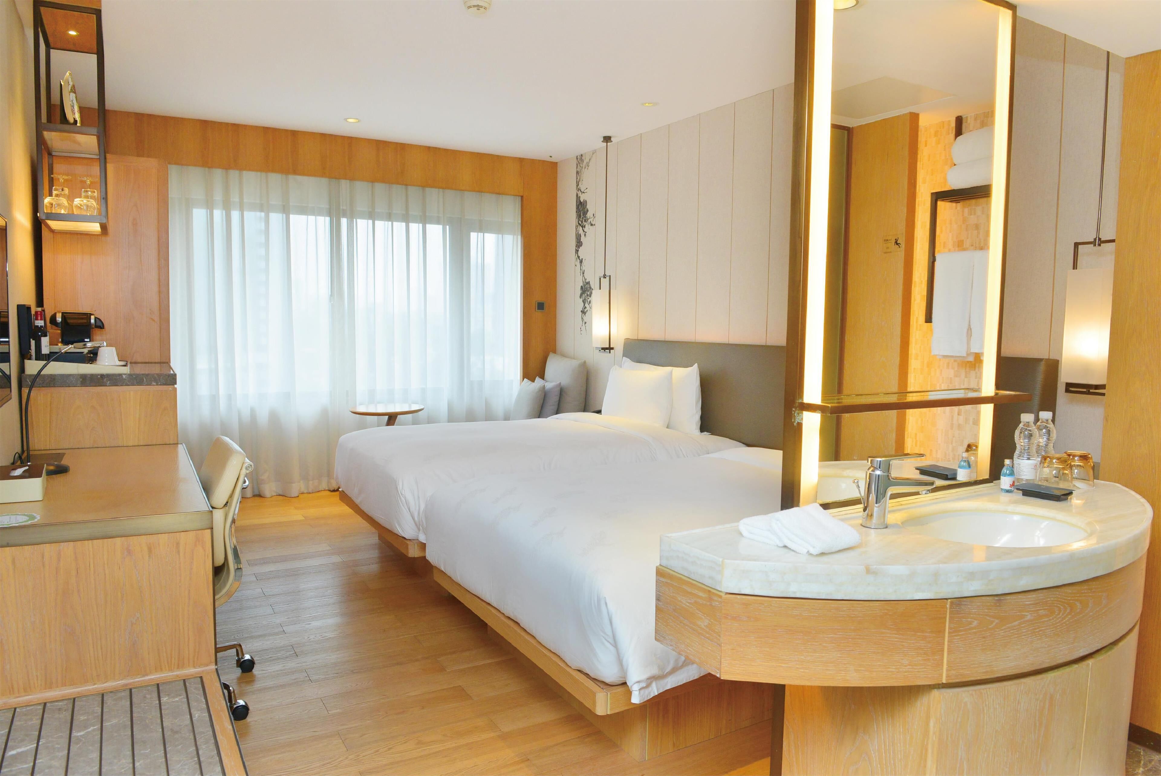 A hotel room with a bed, a desk, a large mirror, a round sink, and a window with curtains.