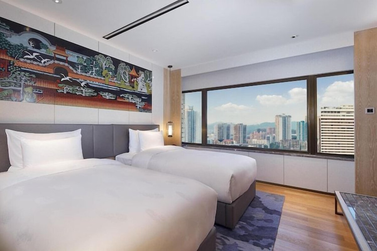 Een hotelkamer met twee bedden, een groot raam met uitzicht op de stad, een schilderij aan de muur en een televisie aan de rechterkant.