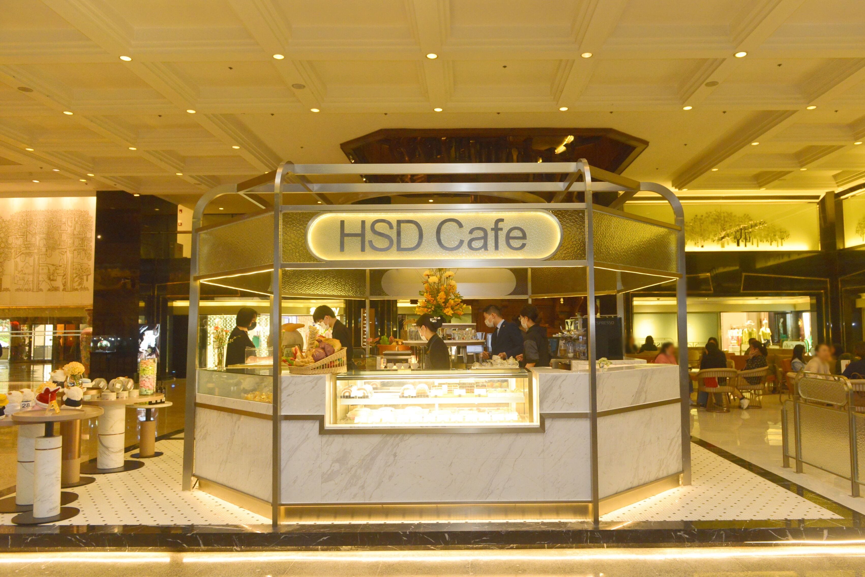 Een HSD Cafe met een marmeren toonbank en een glazen vitrine, gelegen in een winkelcentrum met zitgedeeltes en andere winkels op de achtergrond.