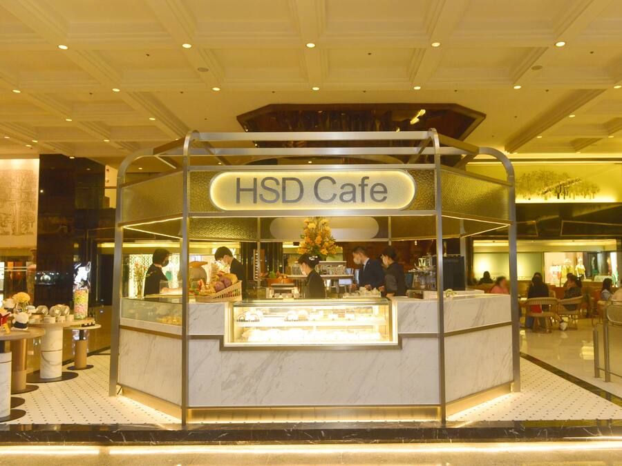 A HSD Cafe with a marble counter and glass display case, located in a mall with seating areas and other shops in the background.