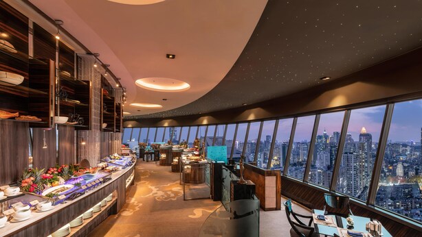 Hotelkamer hoog in een toren met uitzicht over de stad, een ontbijtbuffet met diverse gerechten, en een zithoek met een tafel en stoelen.