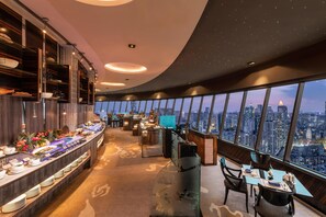 Une chambre d’hôtel située dans un immeuble de grande hauteur, offrant une vue sur la ville, un buffet proposant une variété de plats et un coin salon avec une table et des chaises.