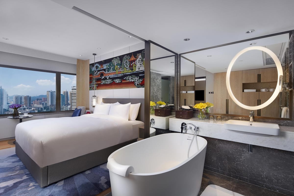 A modern hotel room with a large bed, a bathtub, a large mirror, and a city view.