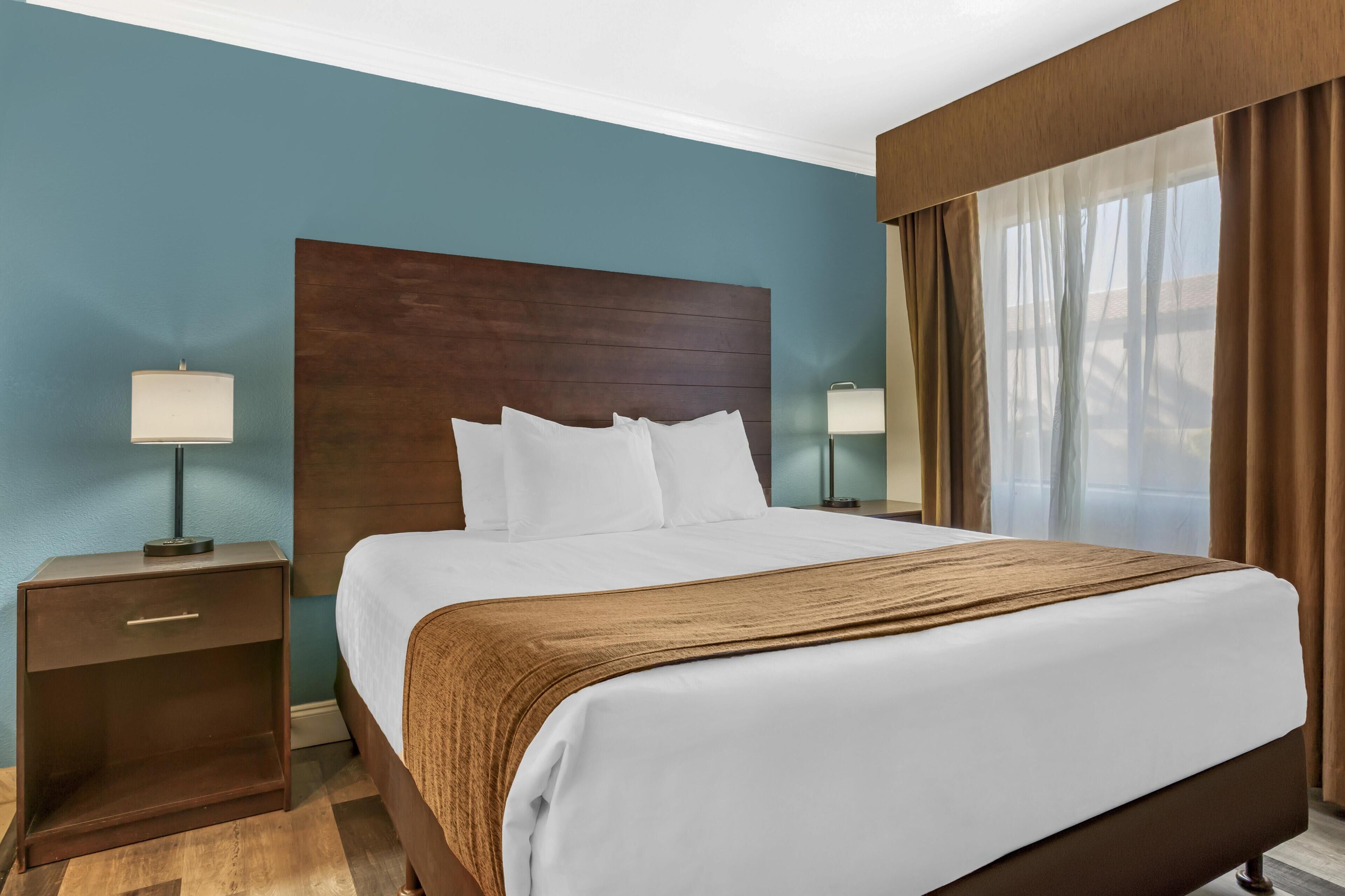 A hotel room with a large bed, two bedside tables, a wooden headboard, and a view of the outdoors through sheer curtains.
