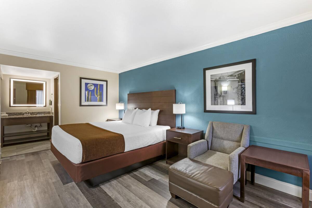 A hotel room with a large bed, a chair, a small table, a nightstand, and a mirror.