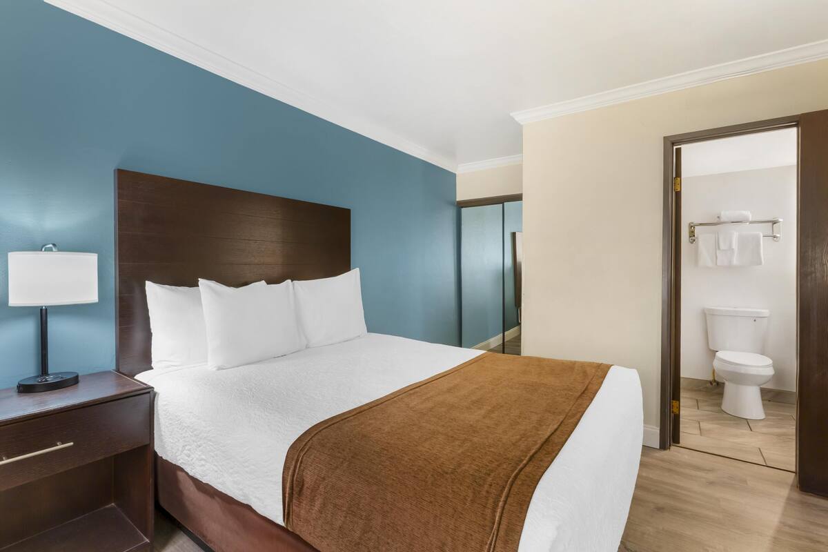 A hotel room with a large bed, a nightstand with a lamp, a wooden headboard, and a view of the bathroom.