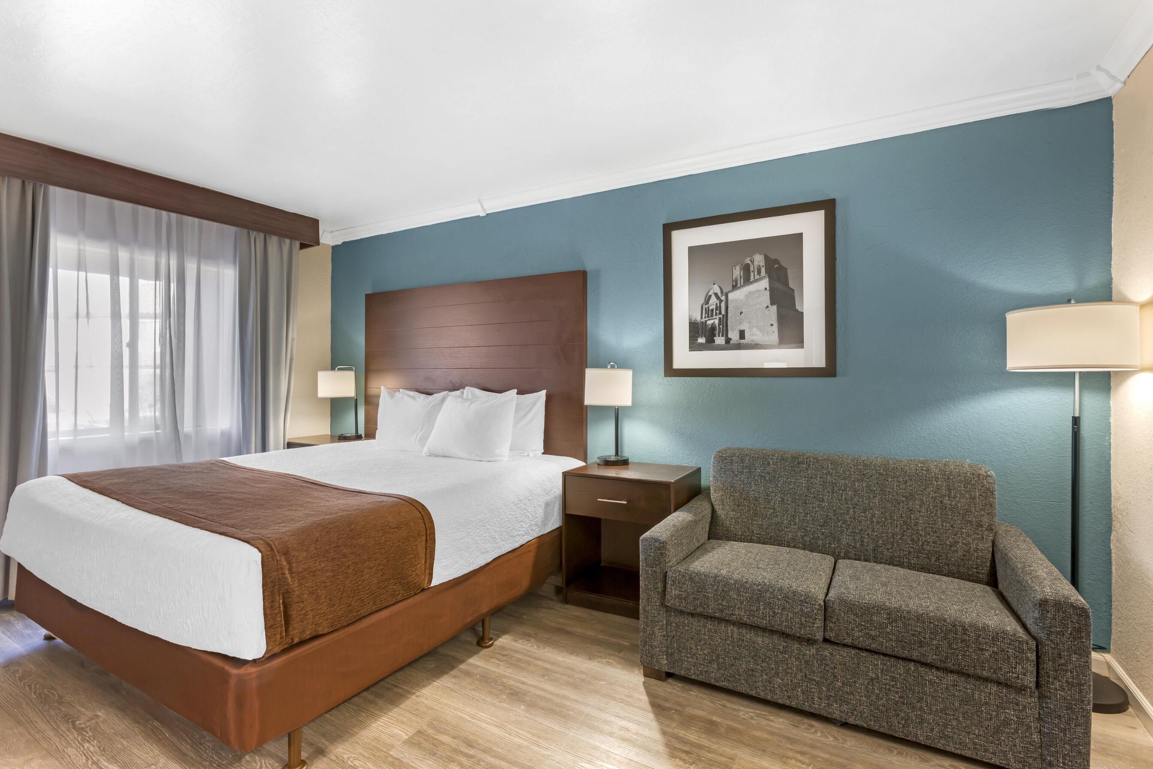 A hotel room with a large bed, a nightstand, a chair, a framed picture on the wall, and a lamp.