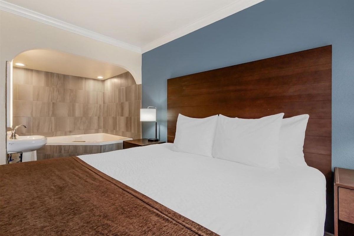 A hotel room with a large bed, a wooden headboard, a bathroom with a bathtub, and a sink.