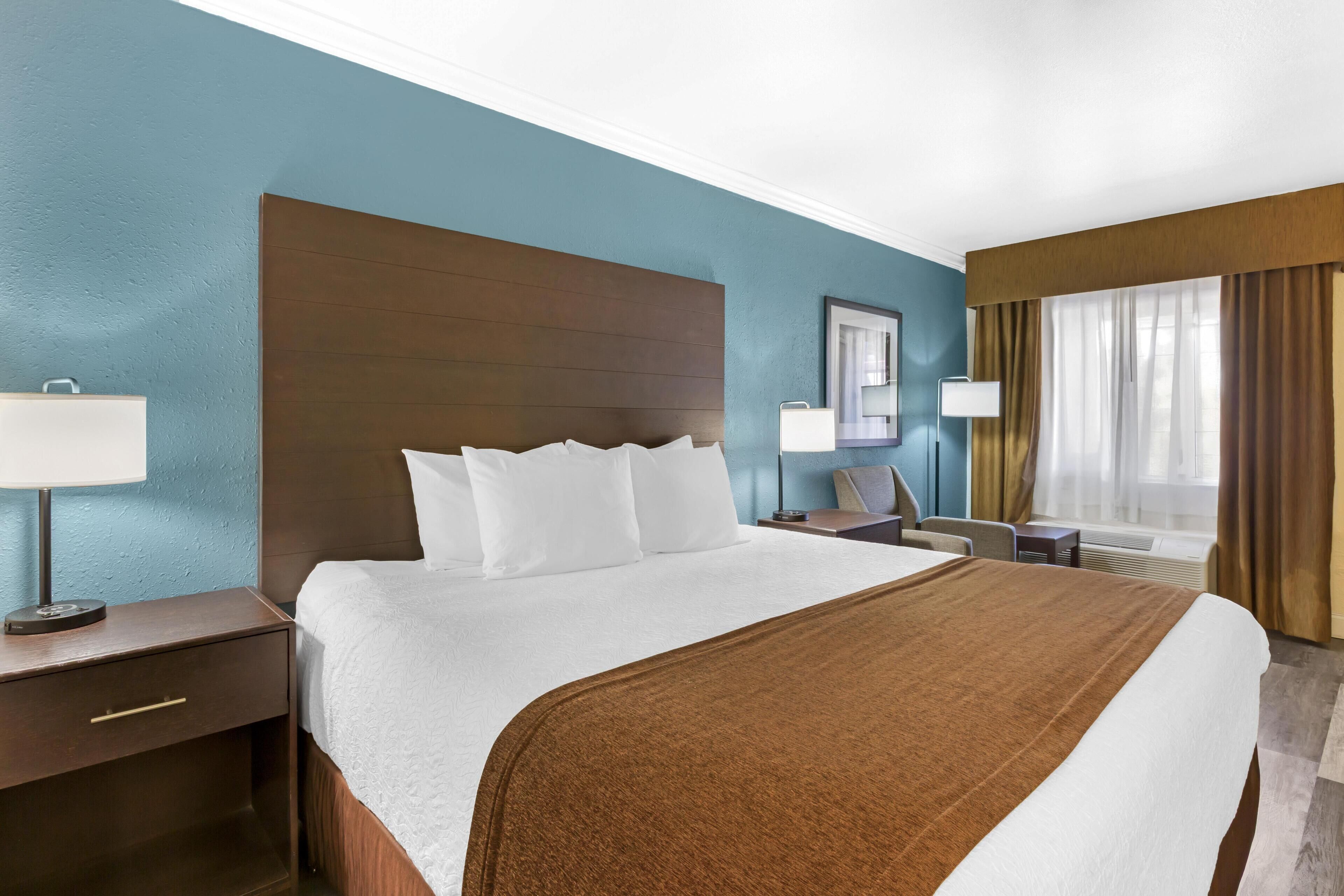 A hotel room with a large bed, a nightstand with a lamp, a desk with a chair, and a window with curtains.