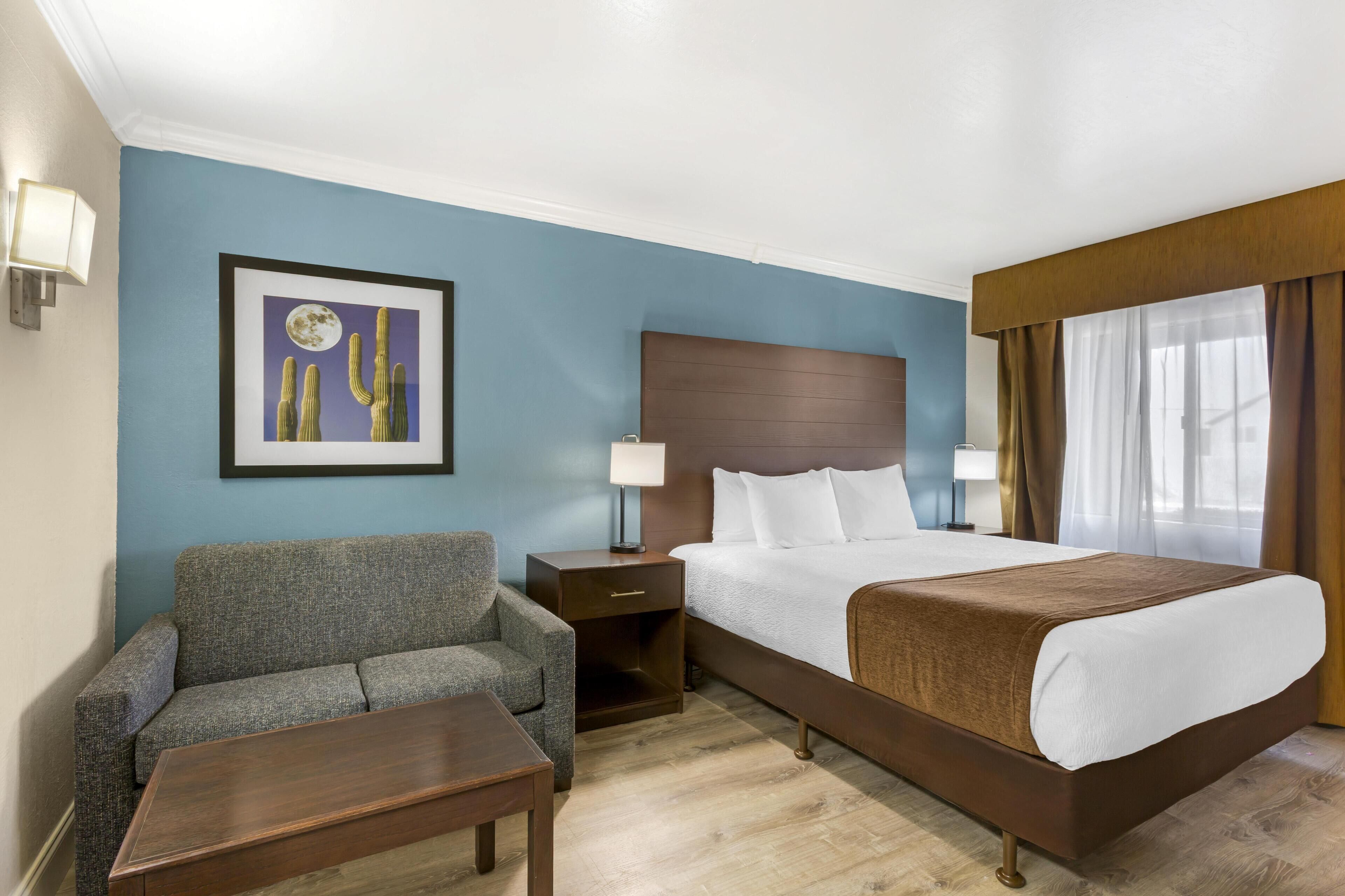 A hotel room with a bed, a sofa, a nightstand with a lamp, and a framed picture on the wall.