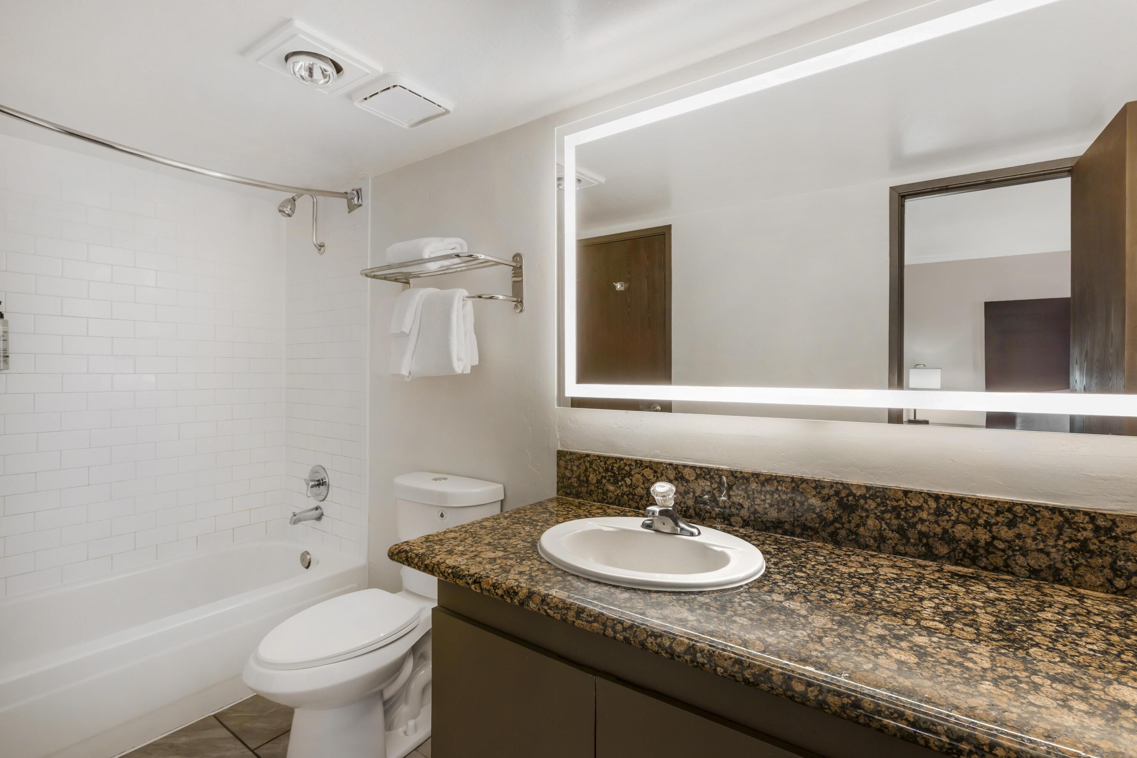 Un baño con inodoro blanco, lavamanos con encimera de granito, ducha con puerta de vidrio y espejo con luz superior.