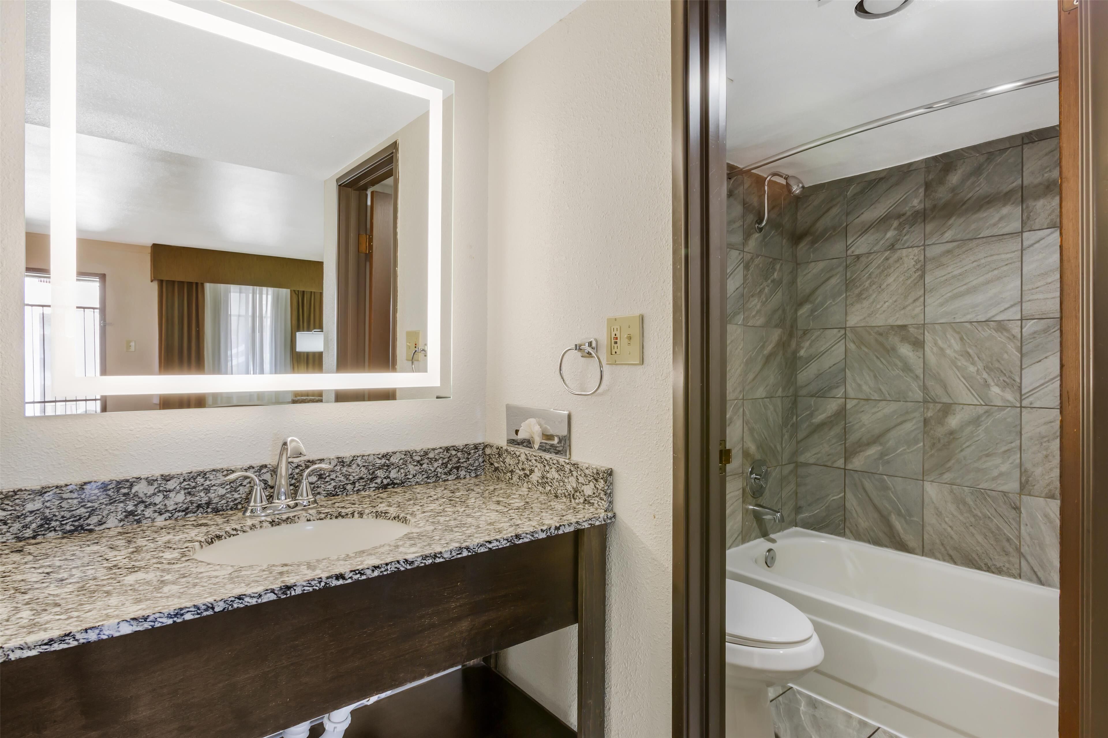 Un baño con lavamanos de encimera de granito, espejo, inodoro y ducha con azulejos de mármol.