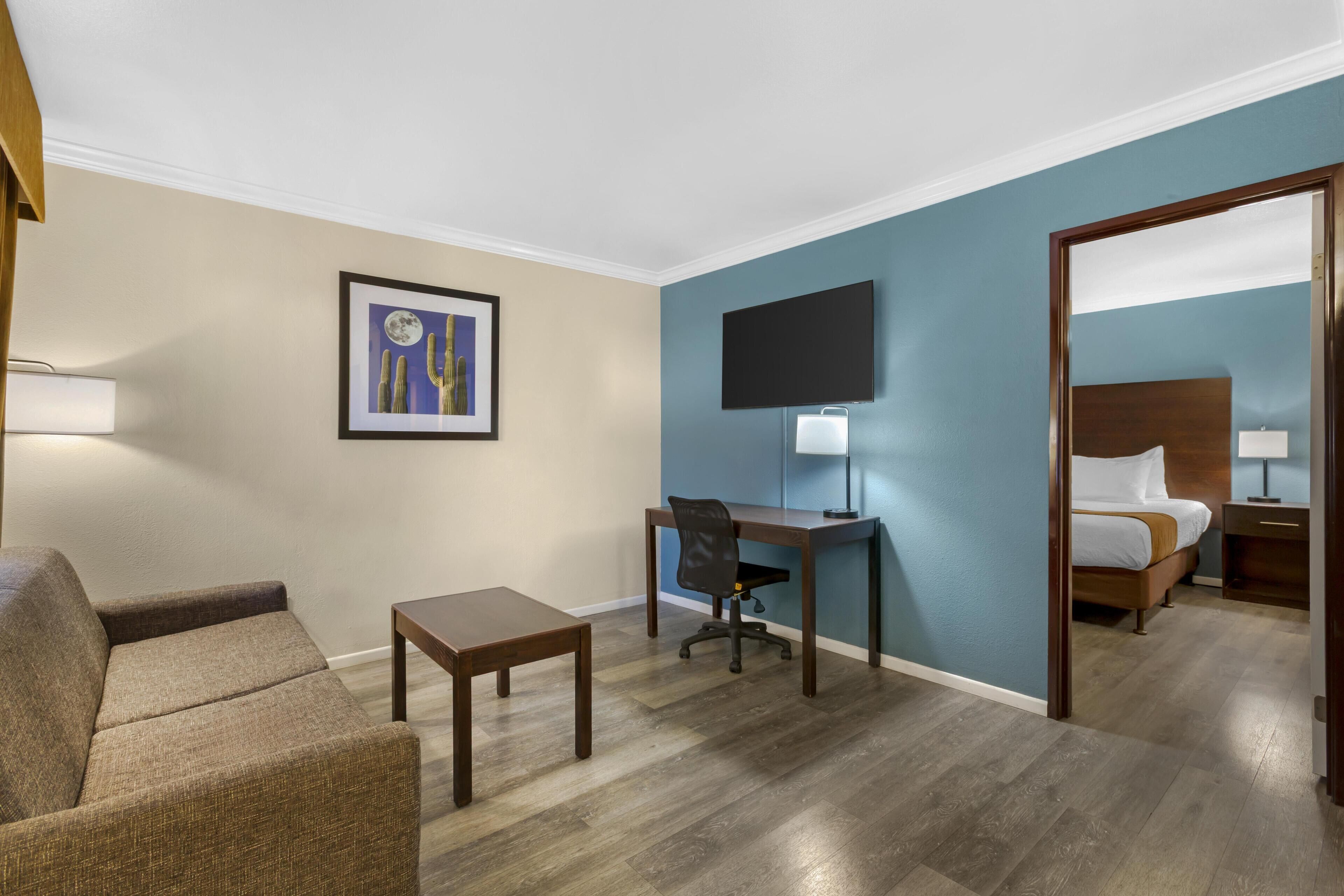 Ein Hotelzimmer mit einem Bett, einem Schreibtisch, einem Sessel, einem kleinen Tisch, einem an der Wand befestigten Fernseher und einem gerahmten Bild.