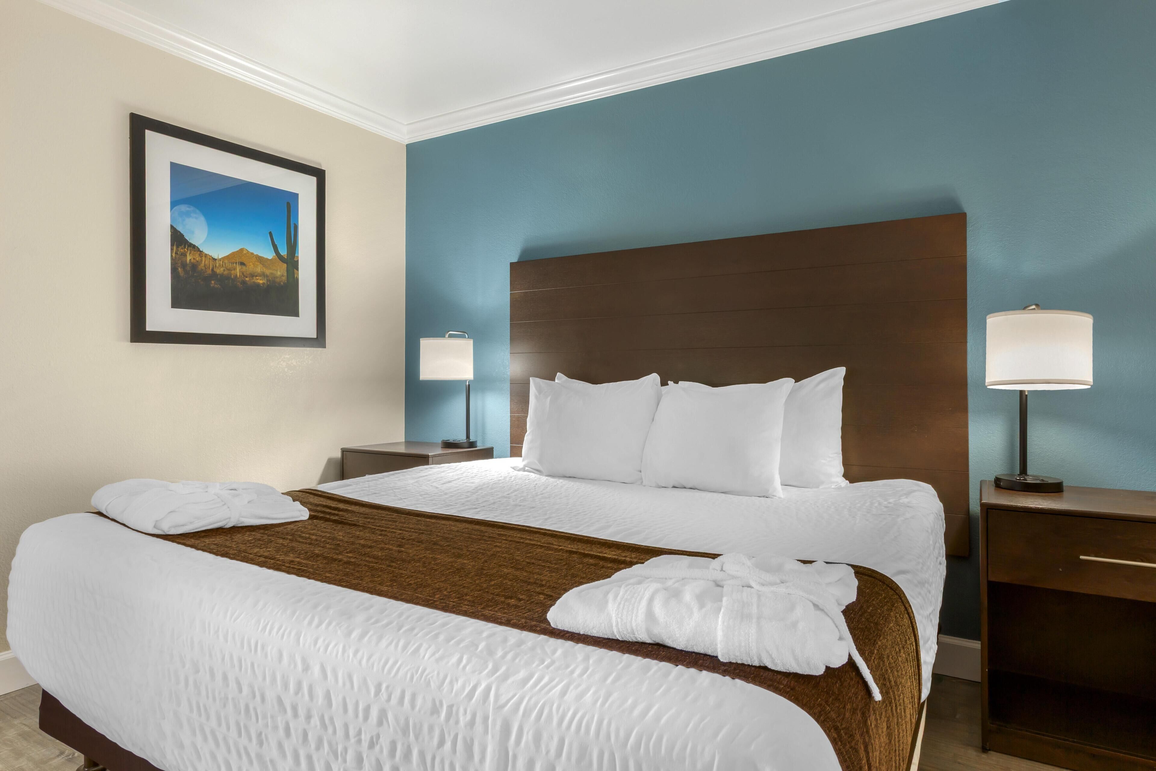 A hotel room with a large bed, two bedside tables, a nightstand, a framed picture on the wall, and two lamps.
