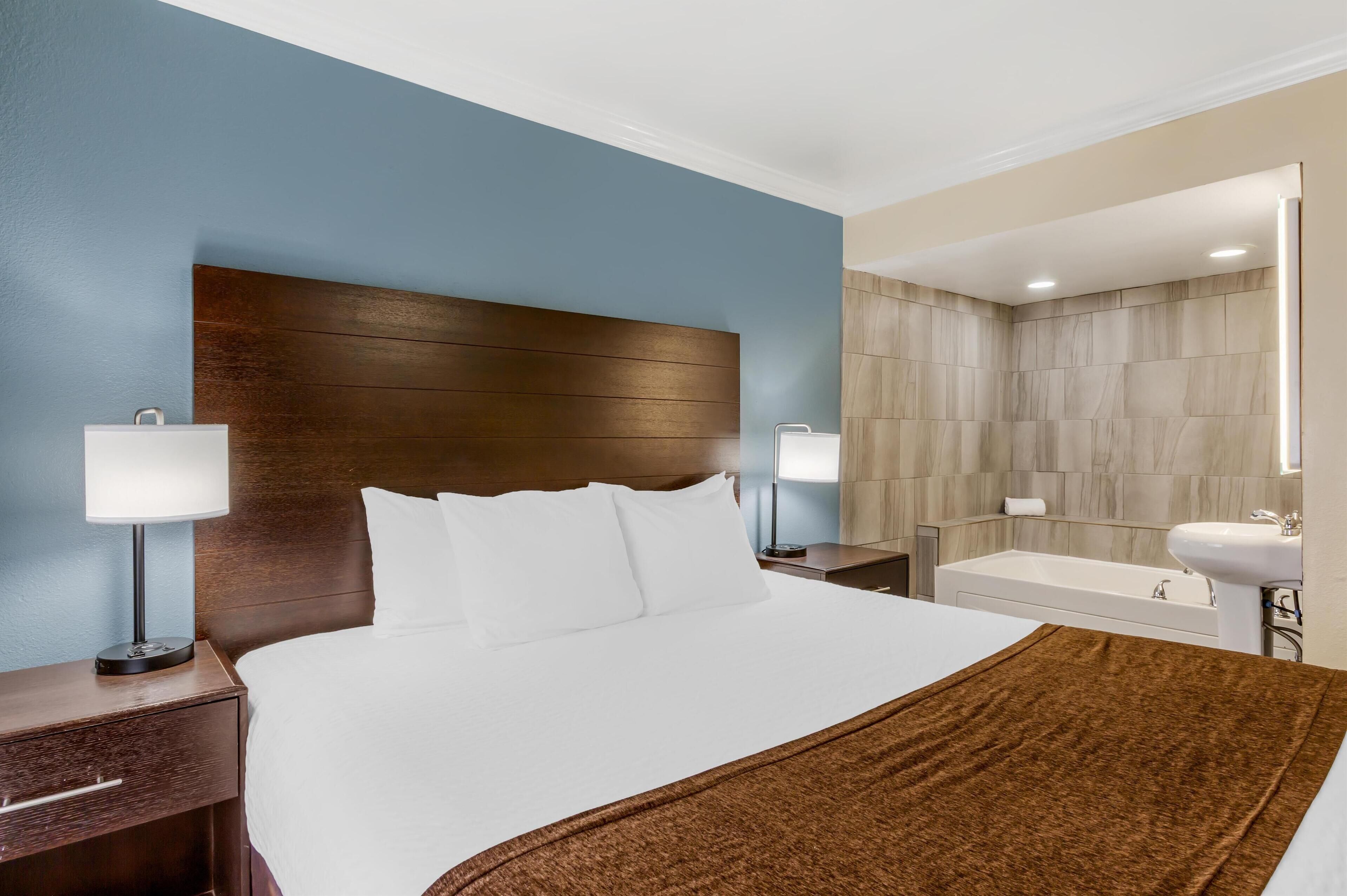 A hotel room with a large bed, two bedside tables with lamps, a brown throw blanket, a bathroom with a bathtub, and a mirror.