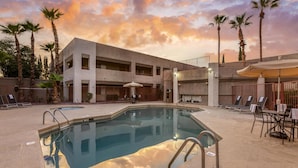 Un área de piscina con un edificio moderno, palmeras y mobiliario exterior.