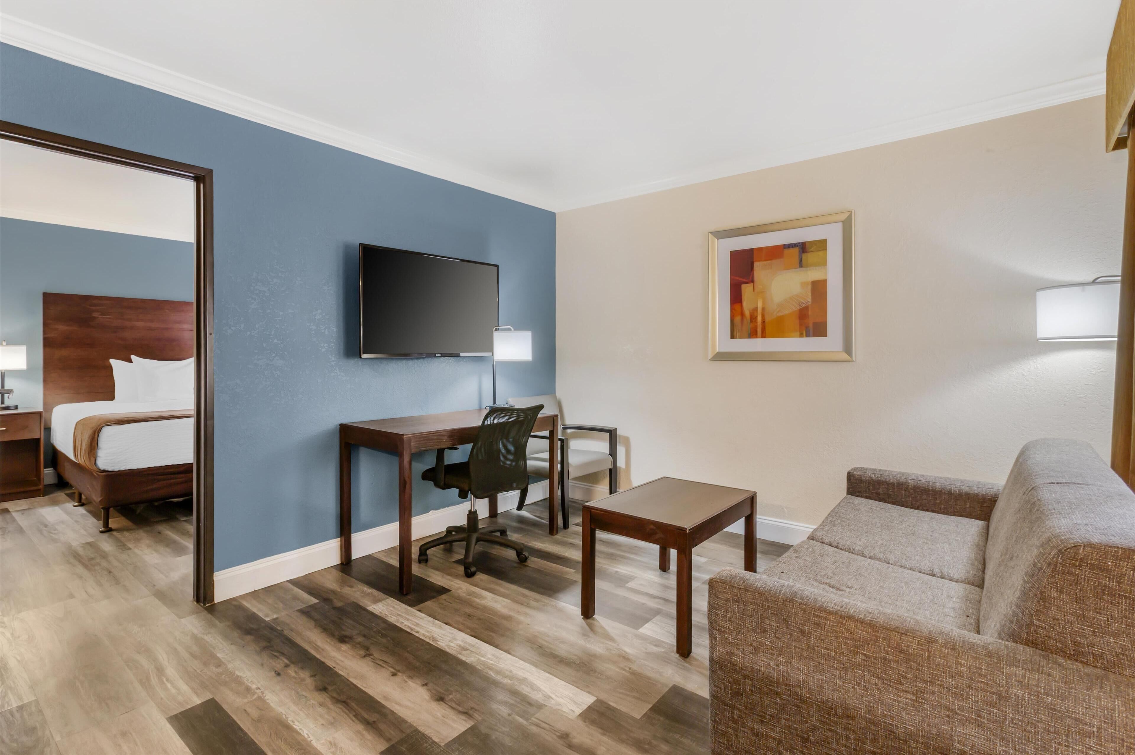 A hotel room with a bed, a desk with a chair, a television, a sofa, and a painting on the wall.