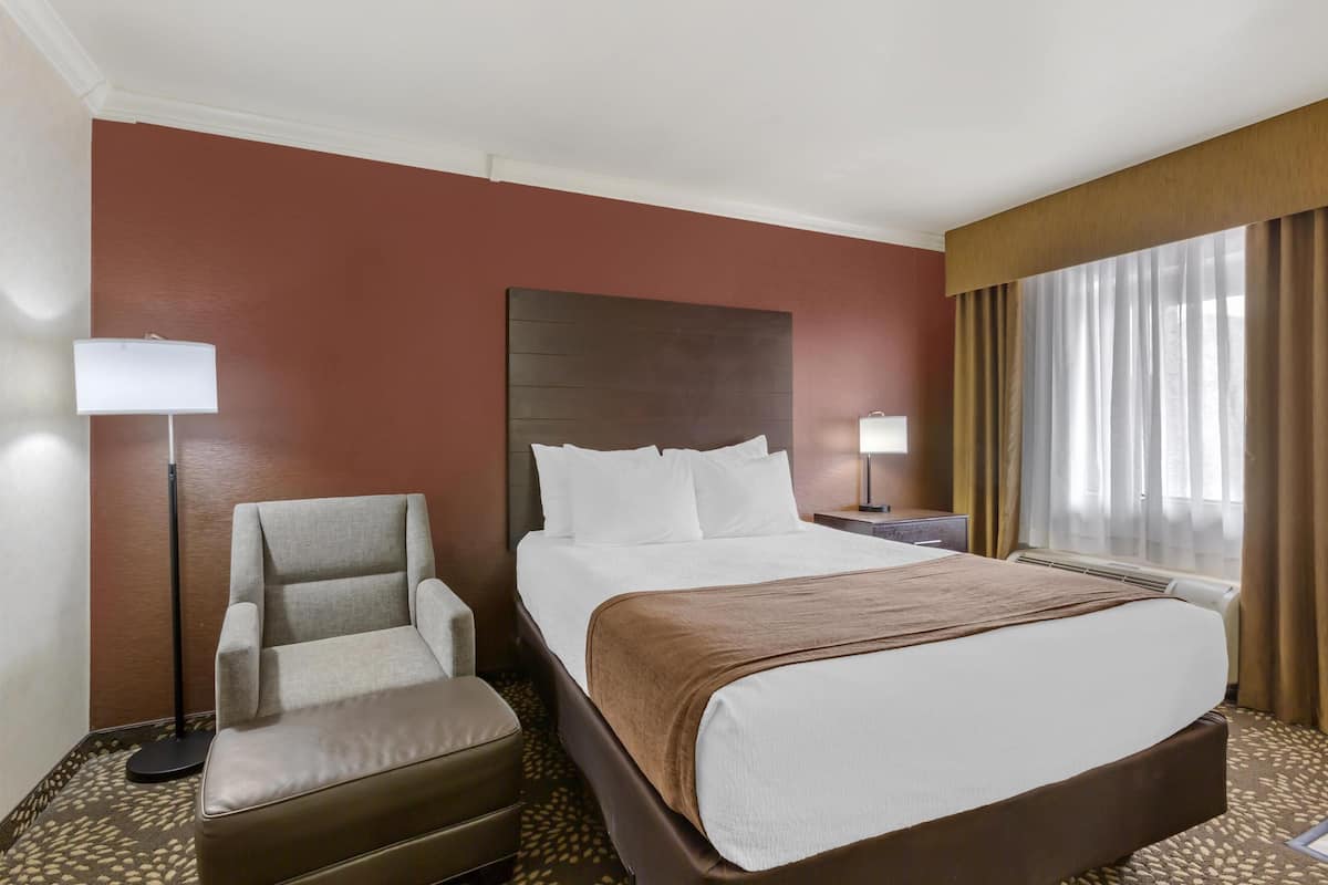 A hotel room with a large bed, a chair, a desk, and a window with curtains.