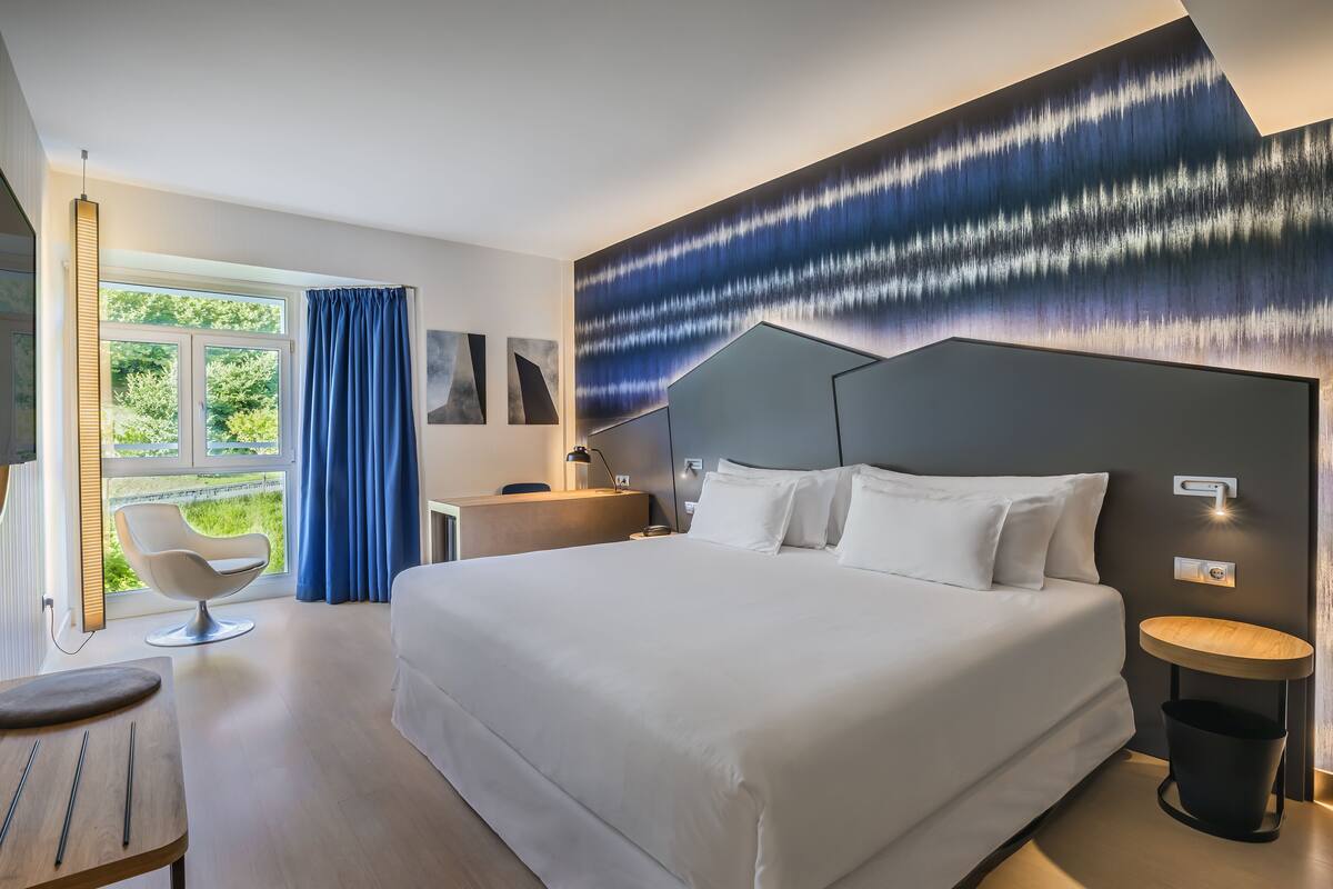 A modern hotel room with a large bed, a desk, a chair, and a view of greenery outside.